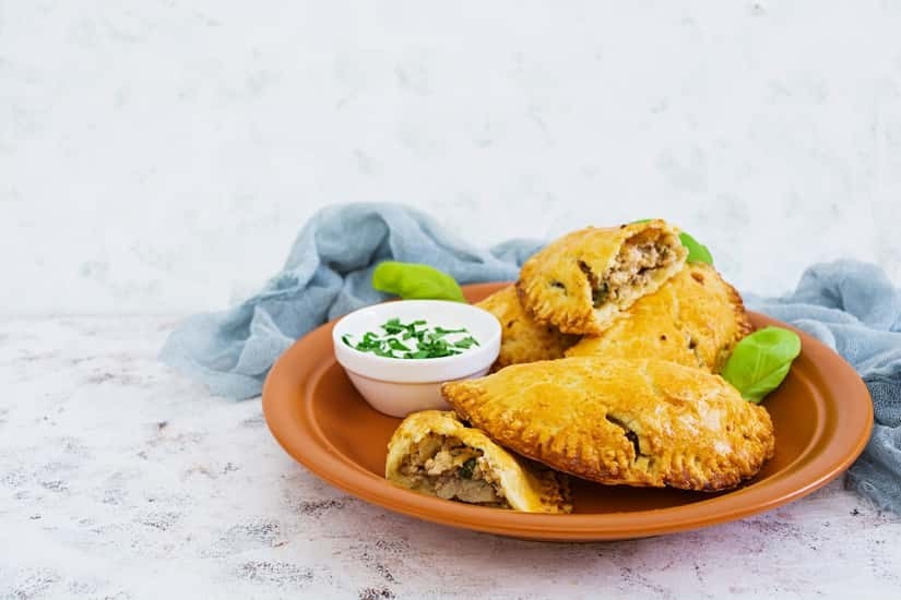 A plate of golden-brown empanadas with a small bowl of dipping sauce garnished with chopped herbs.